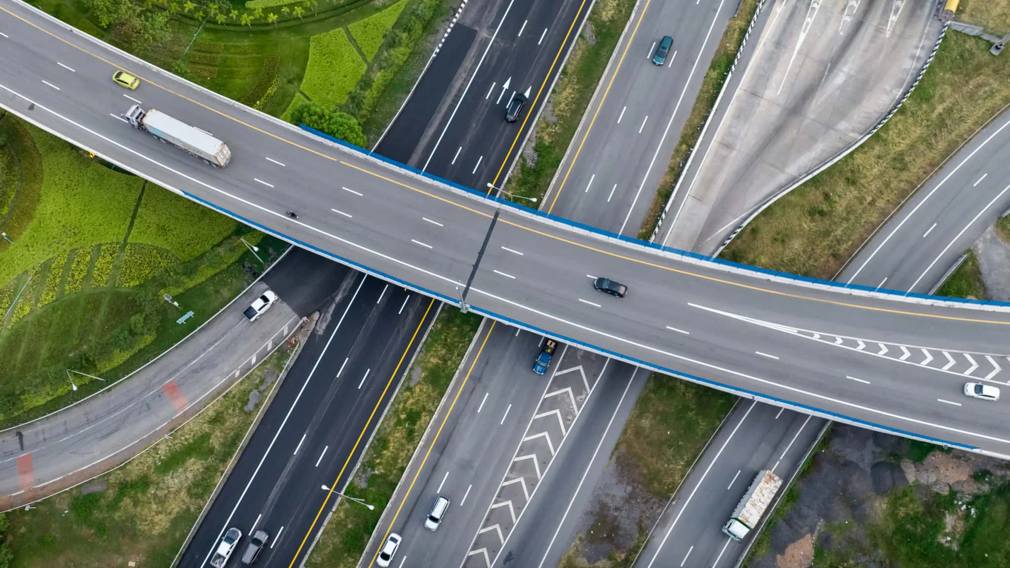 Vista aérea de um trevo rodoviário com caminhões e carros, cercado de vegetação
