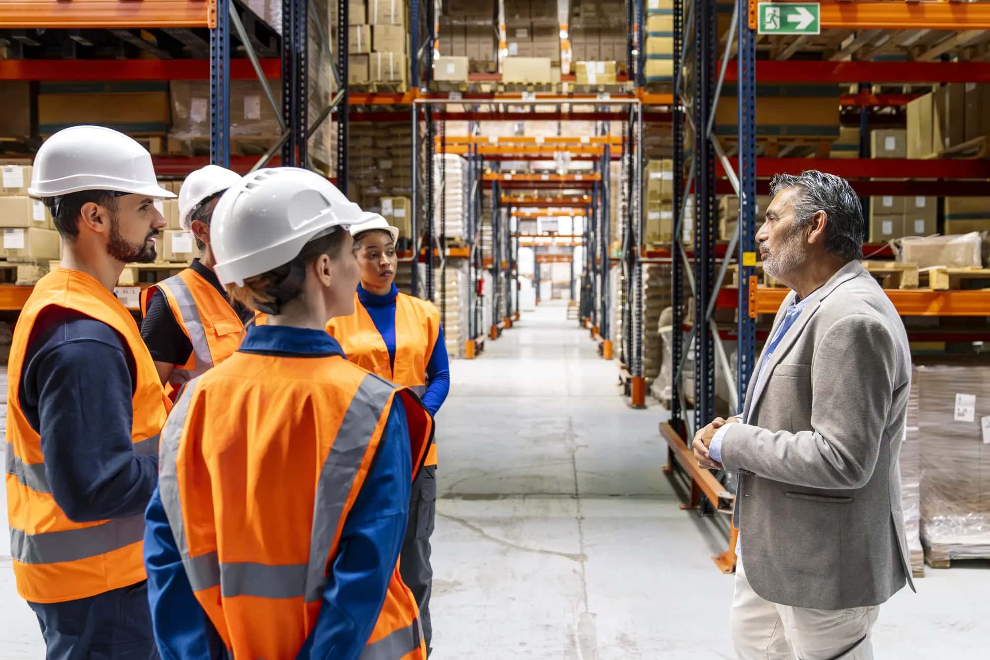 A manager talks with a team in high-visibility vests and hard hats in a warehouse aisle
